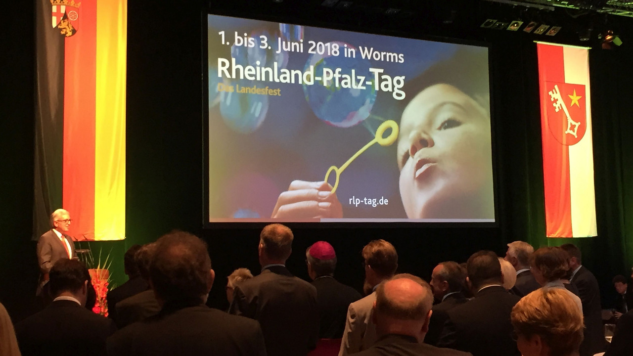 Minister Lewentz hält eine Rede. Auf der Leinwand hinter ihm ist das Motiv des Rheinland-Pfalz-Tages abgebildet.