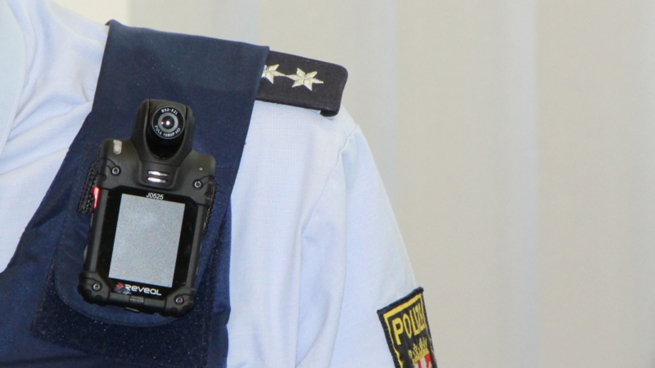 Schulter eines uniformierten Polizisten. An seiner Weste ist trägt er eine Kamera, eine sogenannte Bodycam.