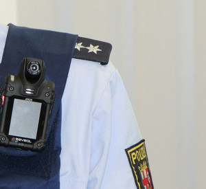 Schulter eines uniformierten Polizisten. An seiner Weste ist trägt er eine Kamera, eine sogenannte Bodycam.