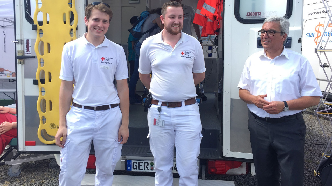 Minister Lewentz mit zwei Mitarbeitern des Deutschen Roten Kreuzes vor einem Rettungswagen.