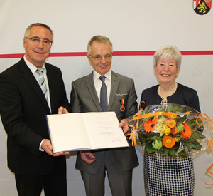Staatssekretär Günter Kern überreicht Werner Schön die Urkunde. Sieglinde Schön hält einen Blumenstrauß
