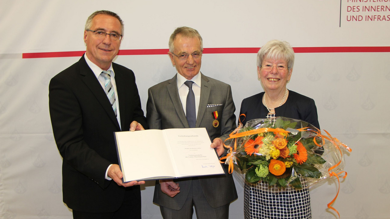 Staatssekretär Günter Kern überreicht Werner Schön die Urkunde. Sieglinde Schön hält einen Blumenstrauß