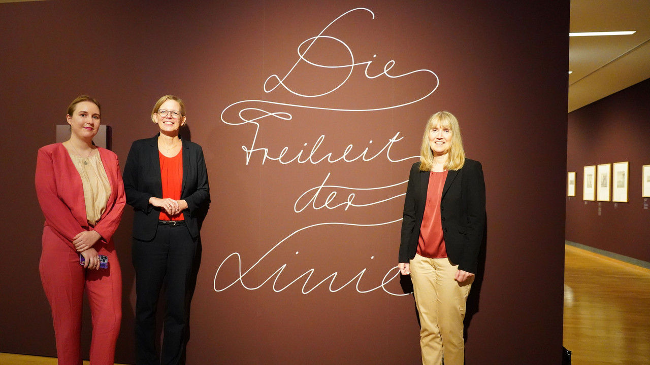 Dr. Maria Aresin, Kuratorin der Ausstellung, Innenstaatssekretärin Simone Schneider und Dr.  Birgit Heide, Direktorin des Landesmuseums, vor dem Logo der Ausstellung. 