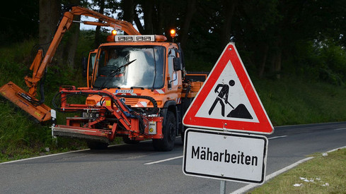 Straßenunterhaltung im Vordergrund steht ein Verkehrsschild mit dem Hinweis Mäharbeit, im Hintergrund ist ein Fahrzeug der Straßenmeisterei zu sehen, dass gerade Mäharbeiten ausführt