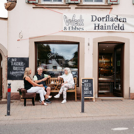 Blick auf den Dorfladen Hainfeld, vor dem fünf Personen an zwei Tischen zusammen sitzen.
