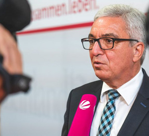 Minister Lewentz spricht in ein Mikrophon, vor ihm steht eine Kamera.