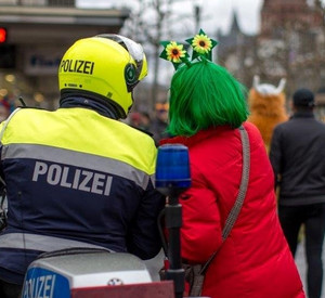 Rückenansicht einer verkleideten Frau, die einen Polizisten auf einem Motorrad etwas fragt.
