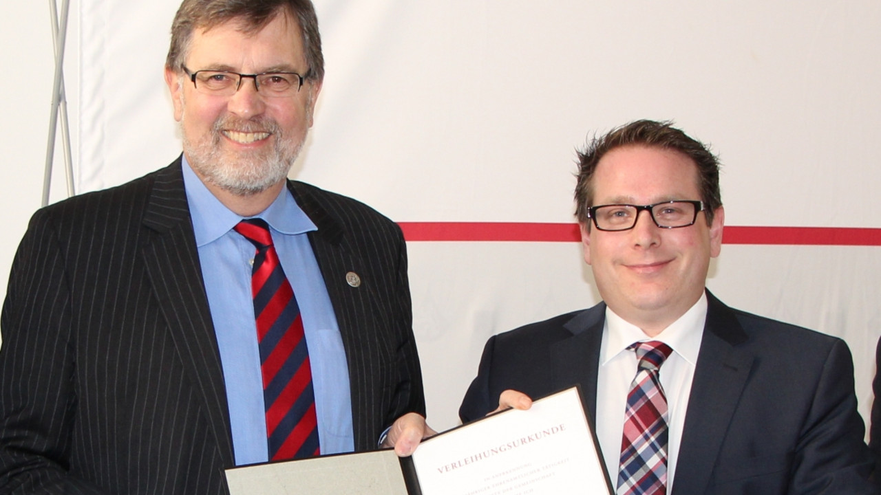Wolfgang Groß und Ministerialdirektor Thomas Linnertz halten gemeinsam die Urkunde über die Verleihung der Ehrenurkunde in die Kamera.