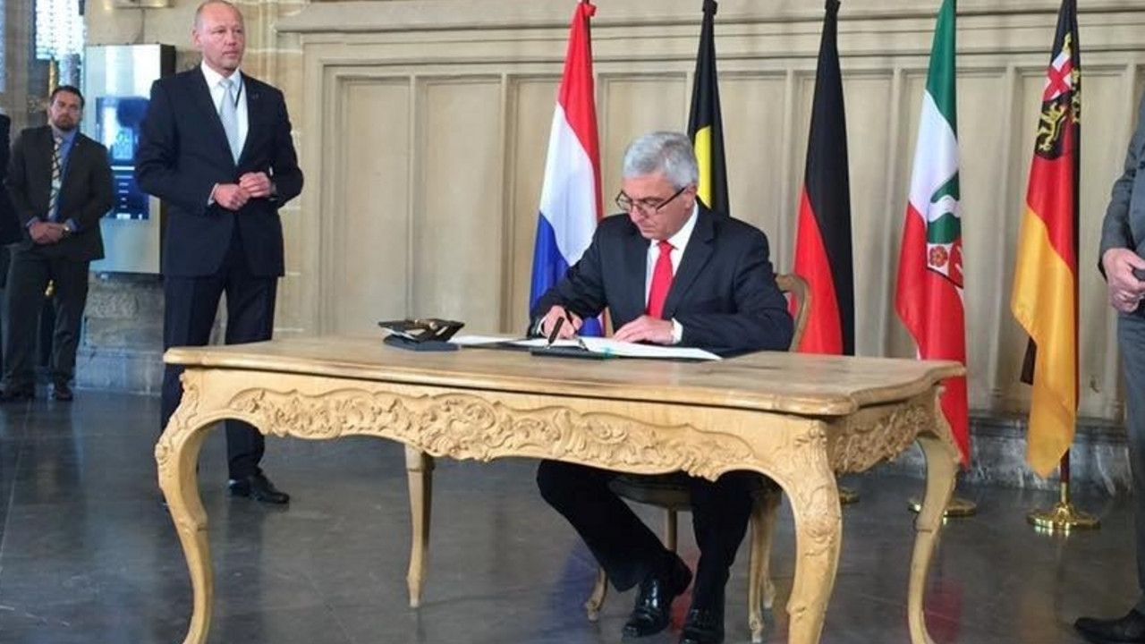 Innenminister Roger Lewentz sitzt an einem antiken Tisch und unterschreibt die Aachener Erklärung. Im Hintergrund sind Flaggen der an der Erklärung beteiligten Länder zu sehen.