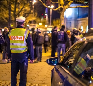 Zwei Polizisten stehen vor einer Gruppe Menschen am Mainzer Hauptbahnhof. Sie haben der Kamera den Rücken zugewandt, einer trägt eine Warnweste mit der Aufschrift "Polizei". Die Abendbeleuchtung ist an. In der rechten Bildhälfte steht ein Polizeiwagen.